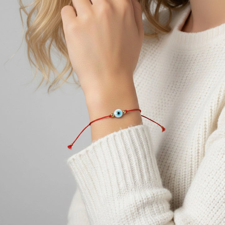 Close-up of a hand wearing a red bracelet with a blue evil eye charm on a light gray background.