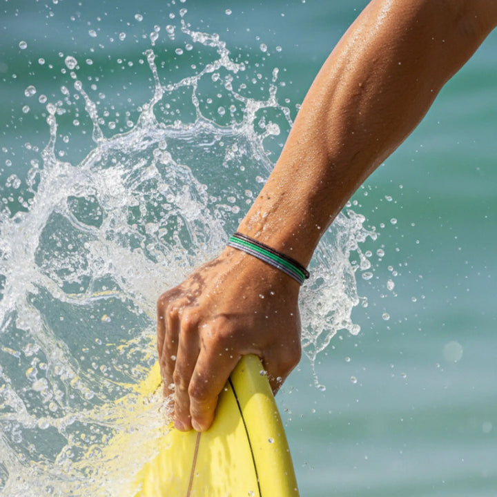 Hand holding a yellow surfboard with water splashing around, wearing a brown, green, and gray bracelet trio against a blurred ocean background