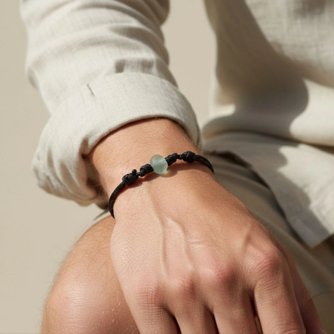 Hand wearing a black cord bracelet with a seafoam glass bead on a neutral background.