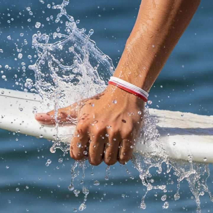 Hand holding a white surfboard with water splashing around, wearing a red, pink, and white bracelet trio, against a blue water background