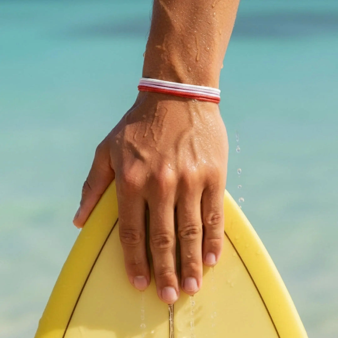 Hand holding a yellow surfboard while wearing a red, pink, and white bracelet trio, with a blurred beach background