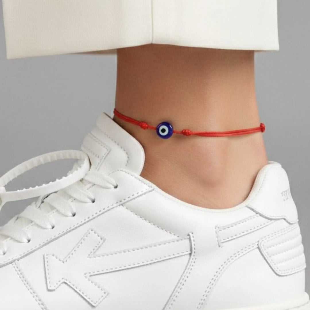 Ankle with a red string bracelet featuring an evil eye charm, wearing a white sneaker.