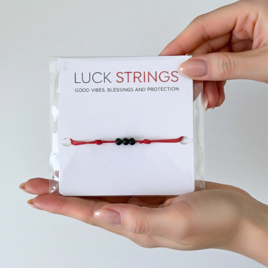 Hand holding a box of 'Luck Strings' with a red cord and black tourmaline bracelet.