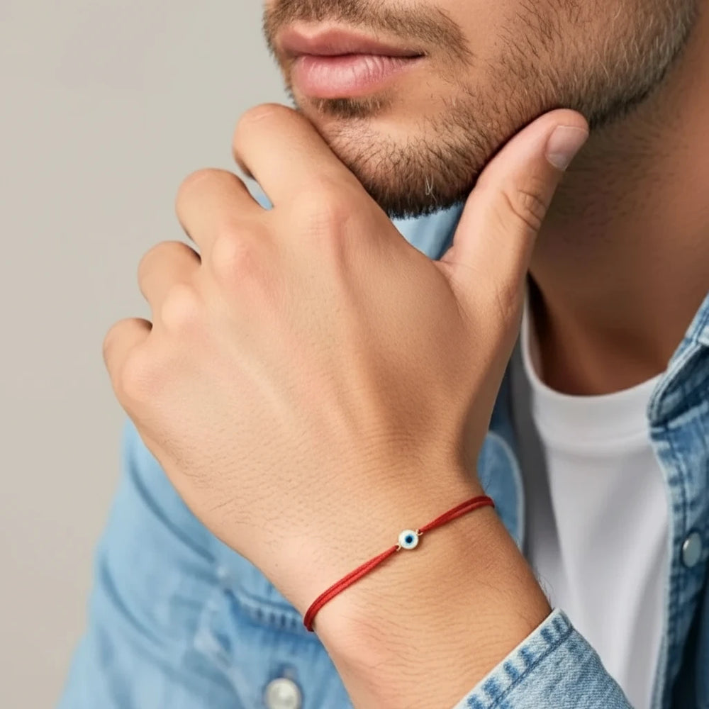 Man wearing a red bracelet with a small gold blue and white evil eye bead, touching his chin.