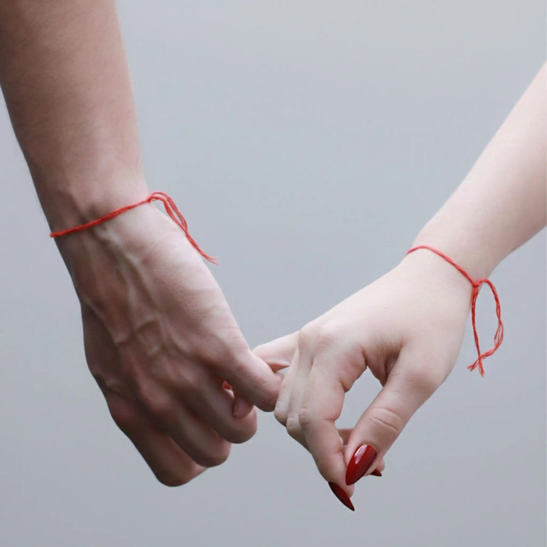 Two hands holding fingers with red strings tied around wrists against a gray background