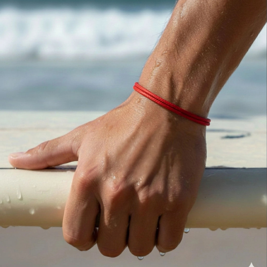 Hand holding a surfboard with a red bracelet on a beach