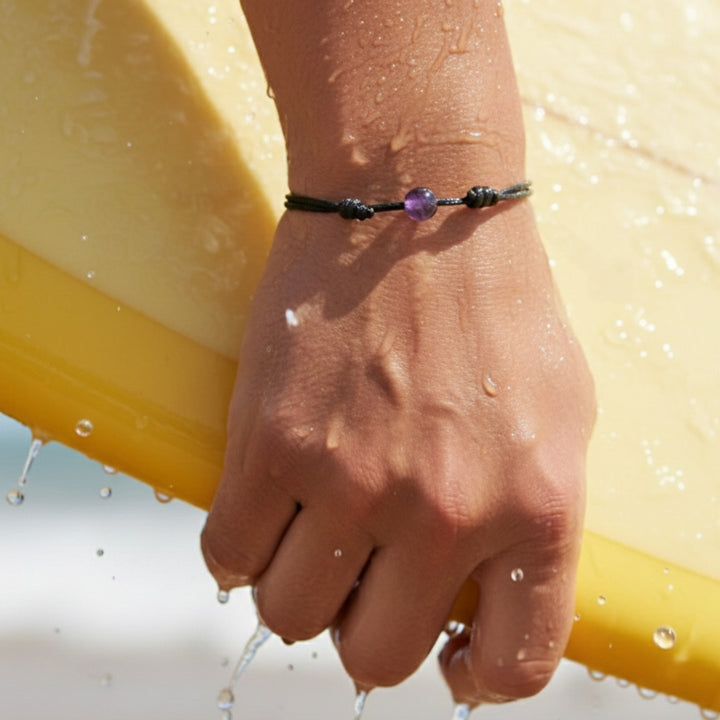 Man wearing smooth amethyst single bead bracelet on black waxed nylon cord while holding a surfboard.

