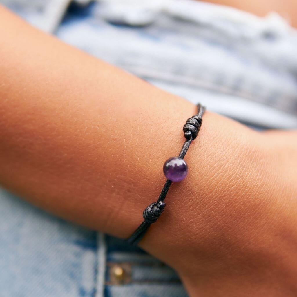 Close-up of smooth amethyst single bead bracelet on black waxed nylon cord.
