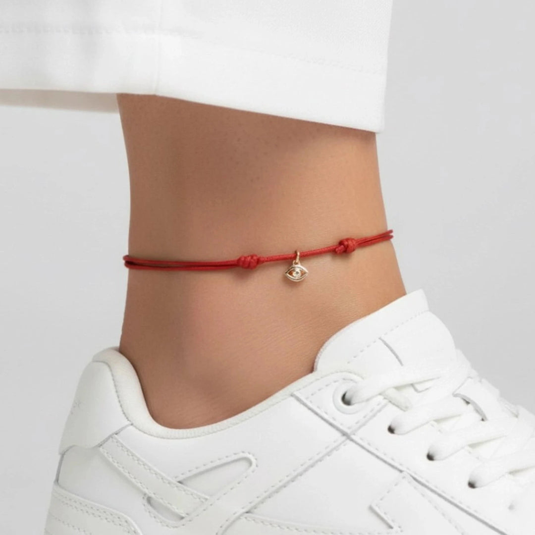 Red anklet with a small charm on a person's ankle, wearing white sneakers against a neutral background