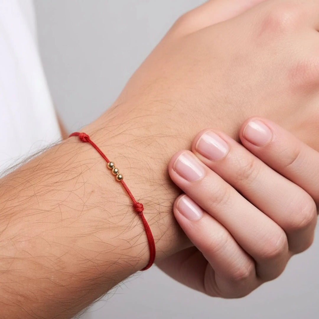 Close-up of a hand wearing a red string bracelet with gold beads on a neutral background