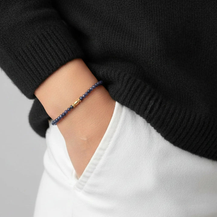 Person wearing a black sweater and white shirt with a blue sapphire beaded bracelet on a neutral background