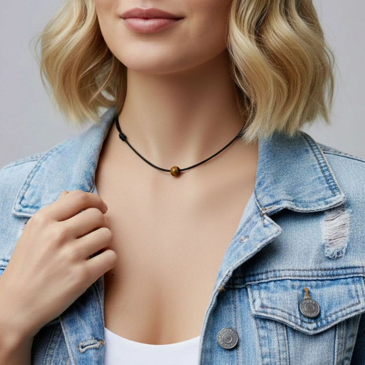 Person wearing a denim jacket and black necklace with a tigers eye brown bead.
