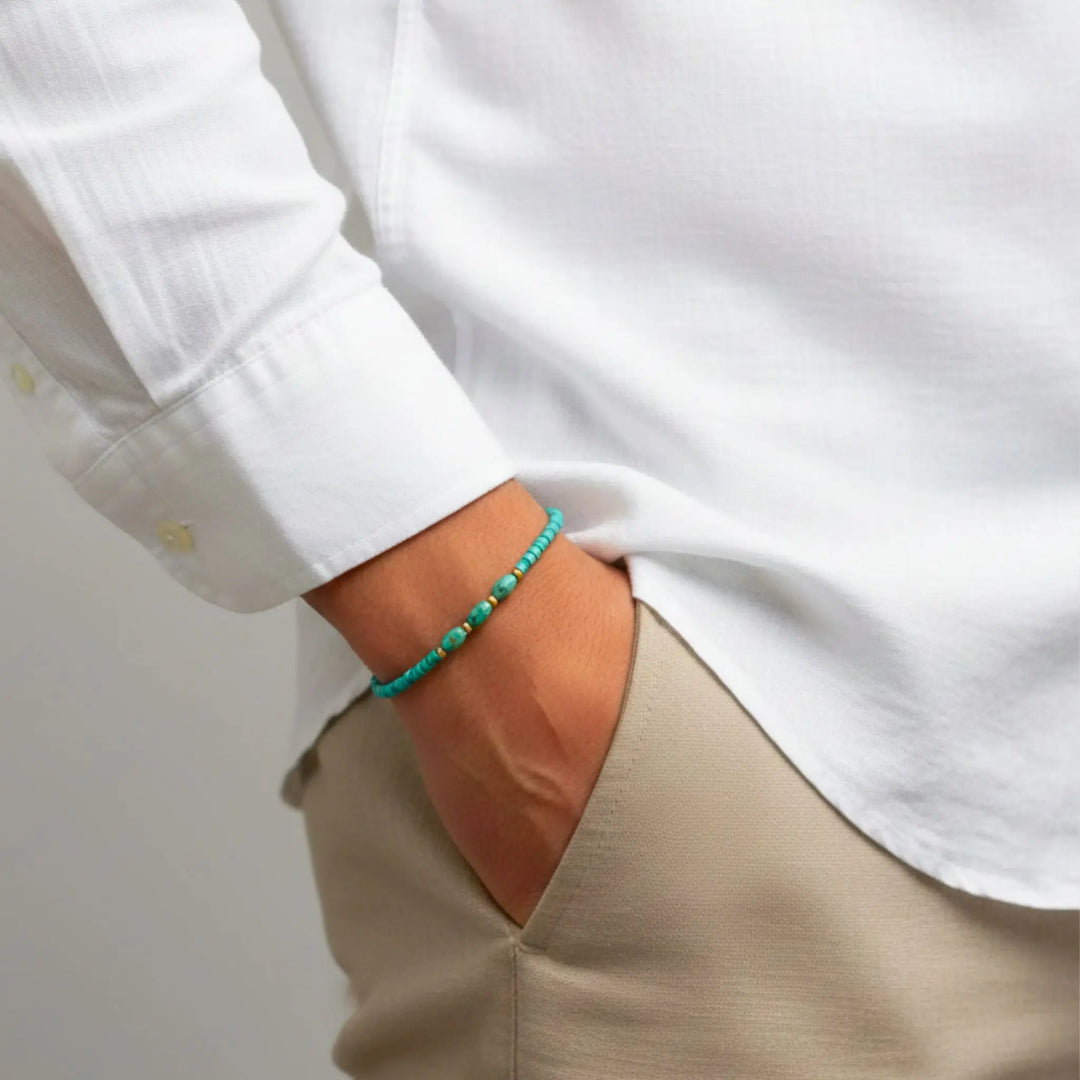 Person wearing a white shirt and beige pants with a turquoise bracelet on a plain background