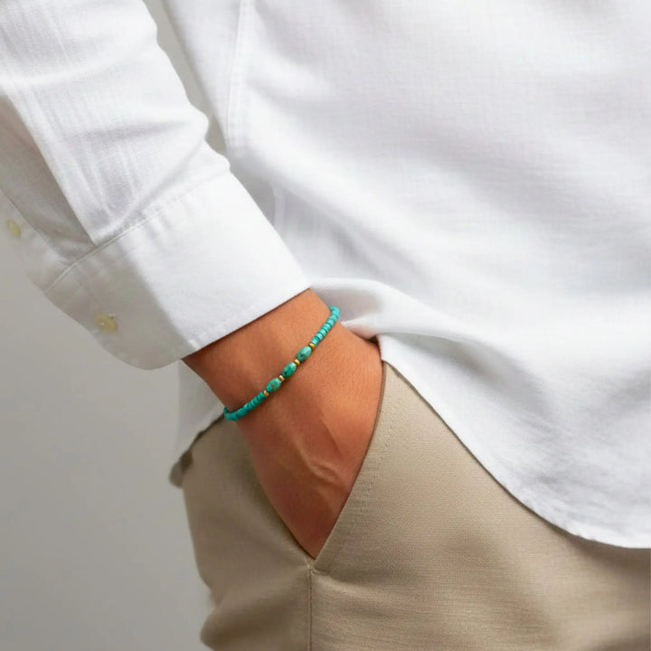 Person wearing a white shirt and beige pants with a turquoise bracelet on a plain background