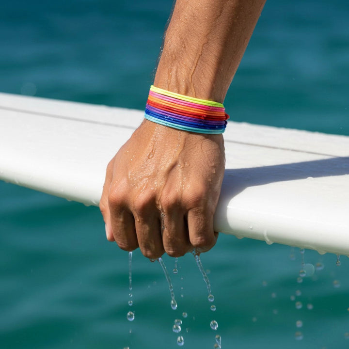 Hand holding a white surfboard with water dripping, wearing a colorful cord bracelet stack, against a blue water background.