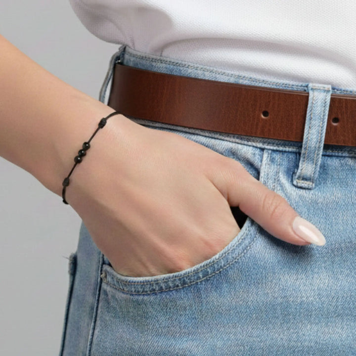 Person wearing a black tourmaline bracelet and a brown belt and blue jeans with a neutral background