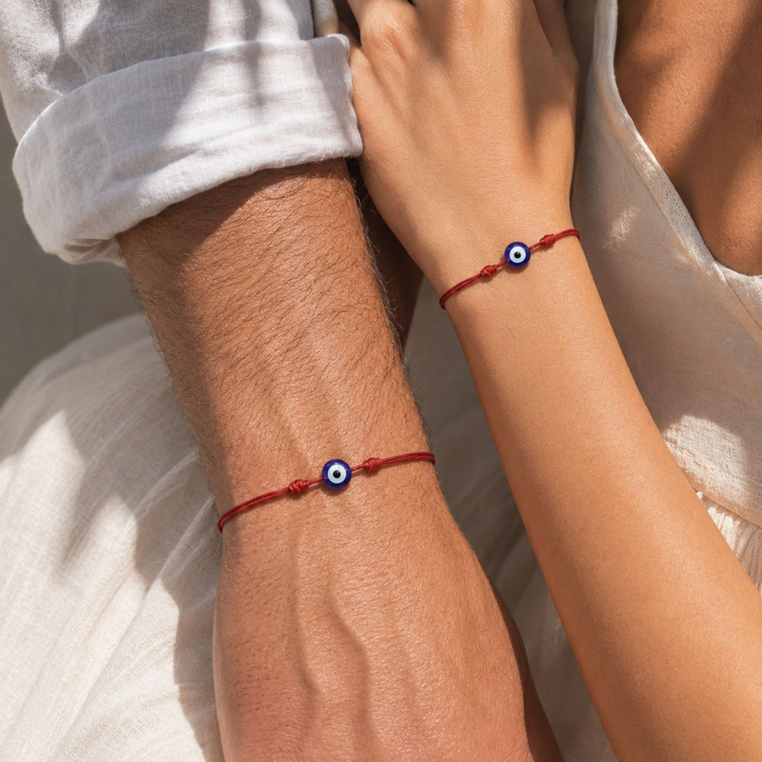 Two red cord bracelets with blue and white evil eye charms worn on a man's and a woman's wrists, styled with white clothing in warm natural light.