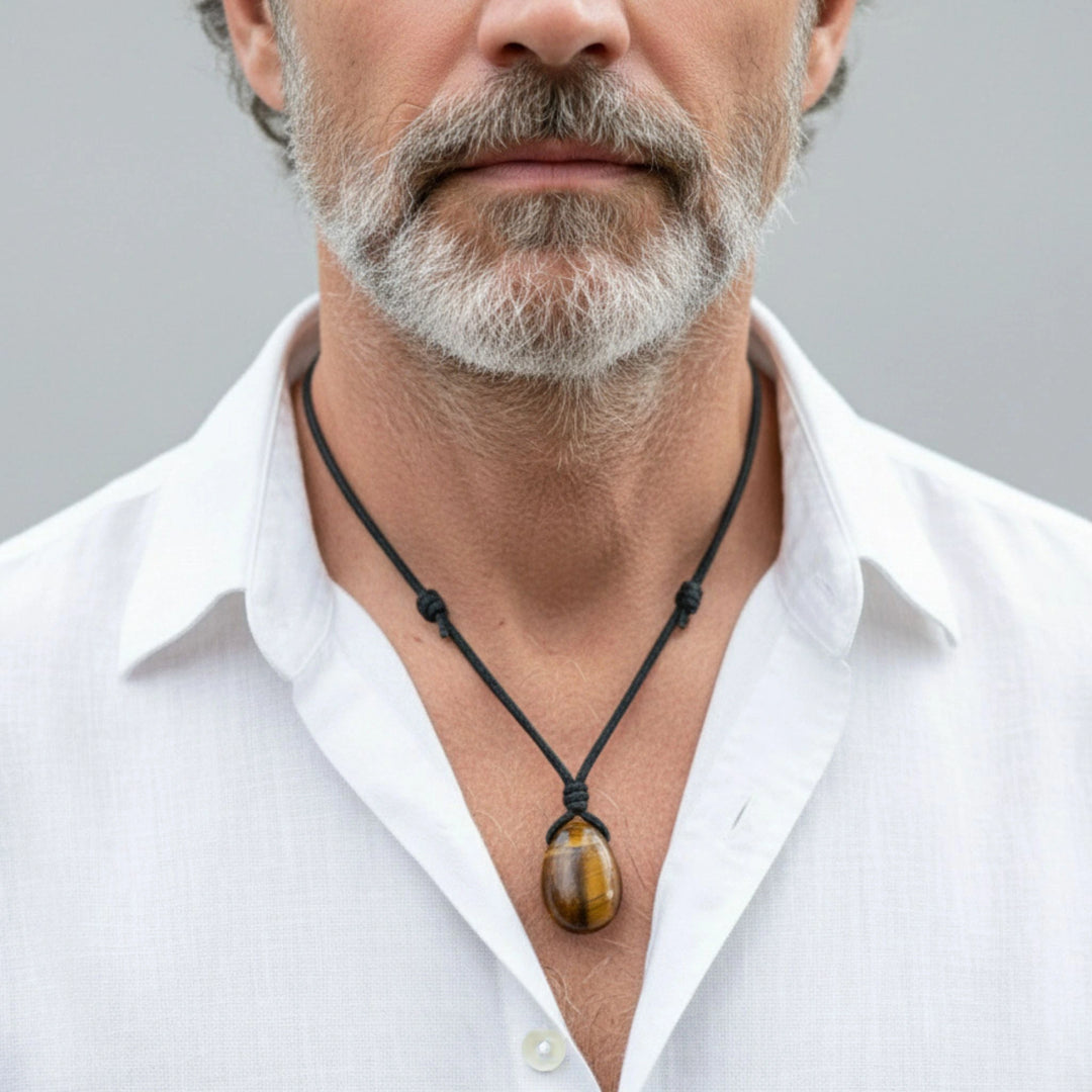 Man wearing a white shirt with a black necklace featuring a tigers eye pendant against a gray background