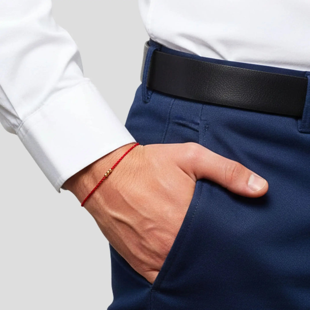 Close-up of a red string bracelet on a person’s wrist, hand in the pocket of navy pants with a black belt and white shirt.