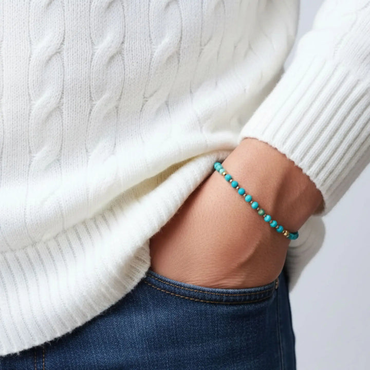 Close-up of a person wearing a white cable-knit sweater and blue jeans with a turquoise beaded bracelet.