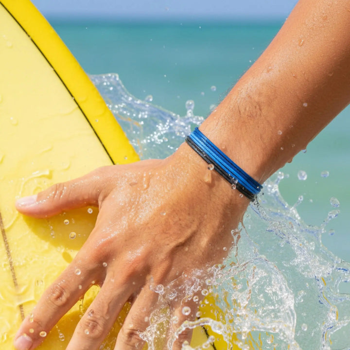 Hand holding a yellow surfboard with water splashing, wearing a blue and black bracelet.