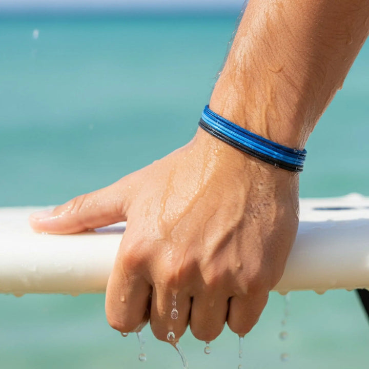 Hand with blue bracelet holding a white surfboard against a blurred beach background