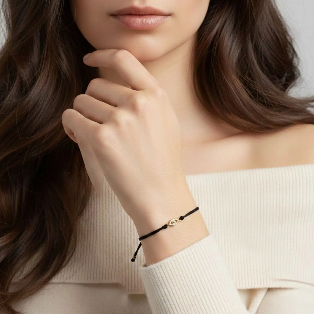 Woman wearing a delicate black bracelet with a gold evil eye gold clasp on a plain background