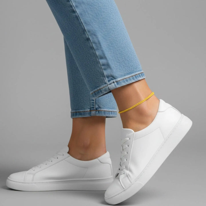 Yellow anklet styled with white sneakers and light blue jeans on a gray background.